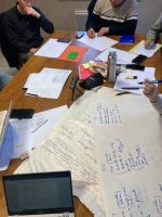 A work table at a meeting. There are papers and a computer, and people around the table, thinking.